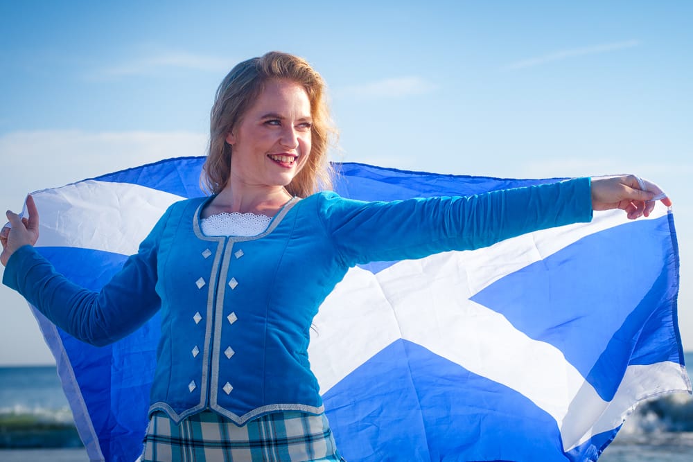 Kasey in her traditional dance attire holds a Scottish flag, blowing in the wind behind her